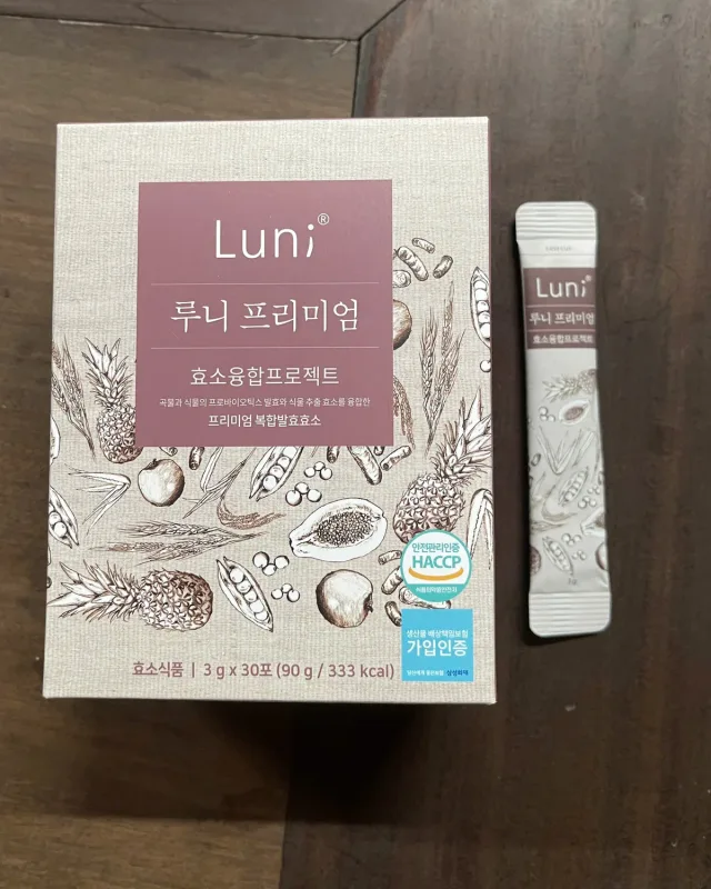 Hộp Luni Premium 90 g và một gói 3 g đặt cạnh nhau trên mặt bàn gỗ sẫm màu, nhìn từ trên xuống